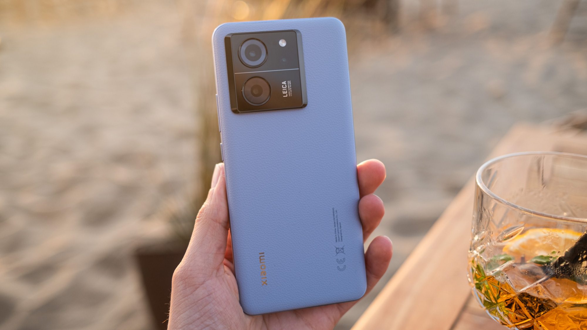 Xiaomi 13T Pro Smartphone mit blauer Rückseite aus Kunstleder vor Strand und Tisch mit Aperitiv.