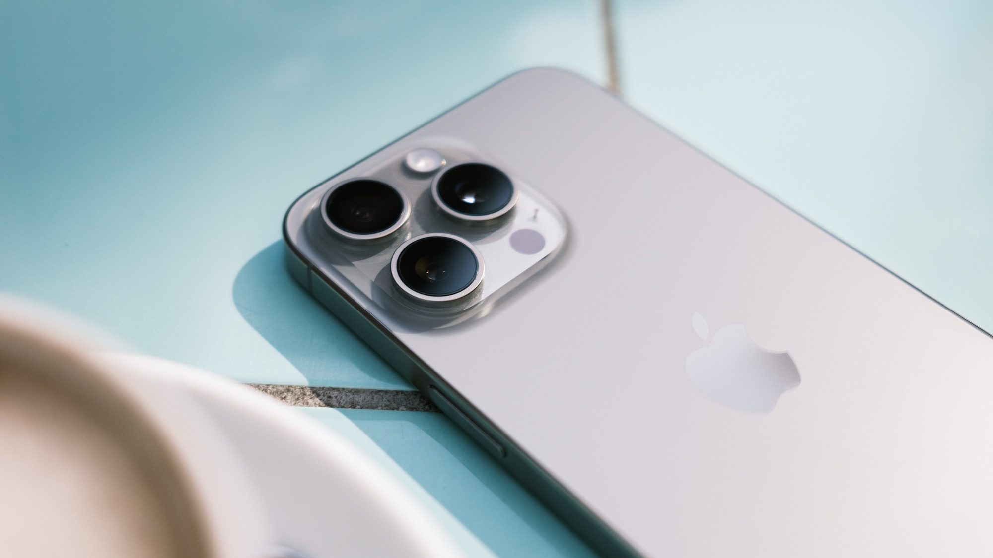Triple-Kamera des Apple iPhone 15 Pro Max auf blauen Fließen liegend.