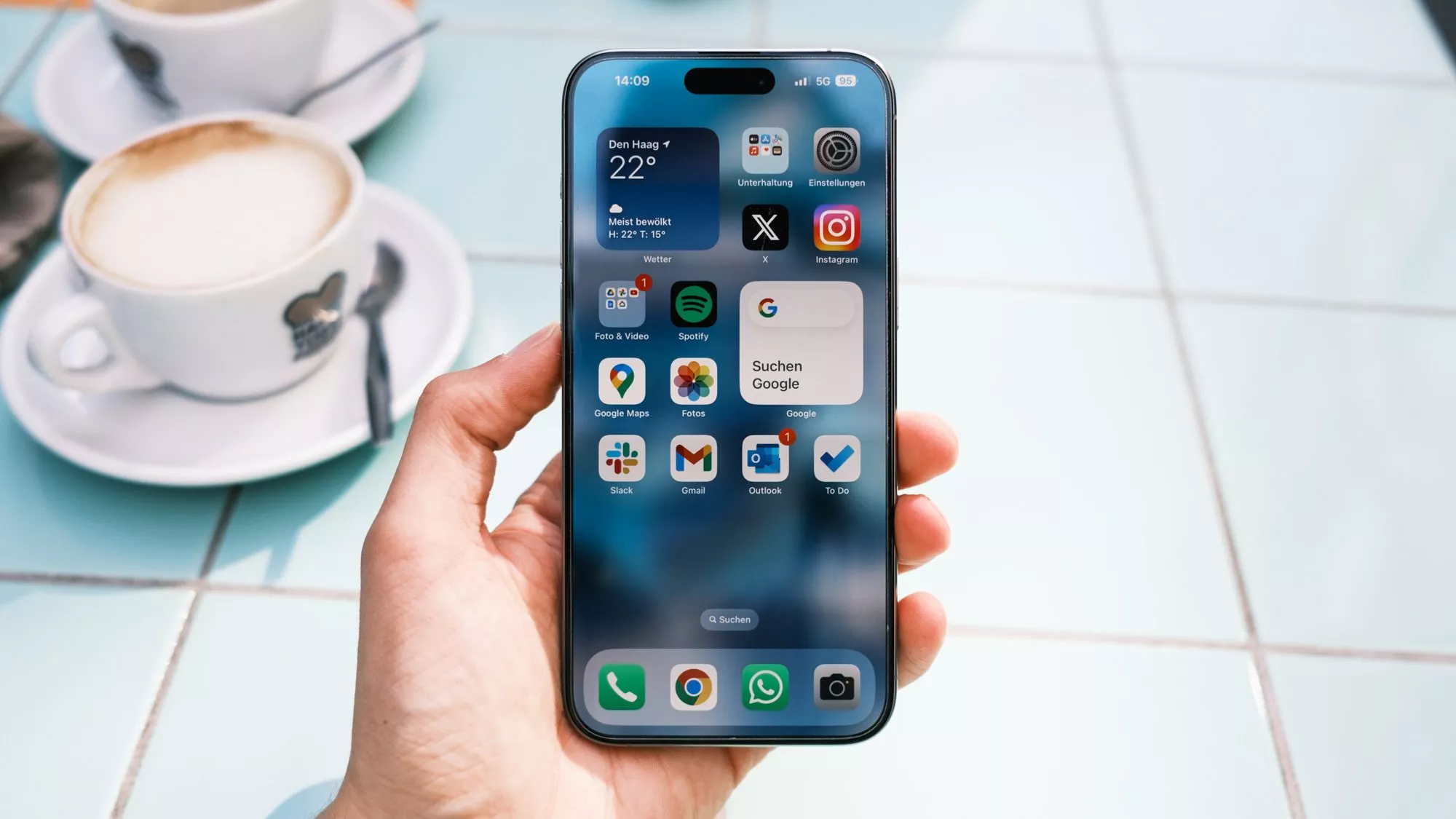 Apple iPhone 15 Pro Max in der Hand mit blauen Fließen und Kaffee im Hintergrund.